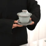 Žmogus laiko keraminį gaiwan indelį su apsauginiais kraštais nuo karščio